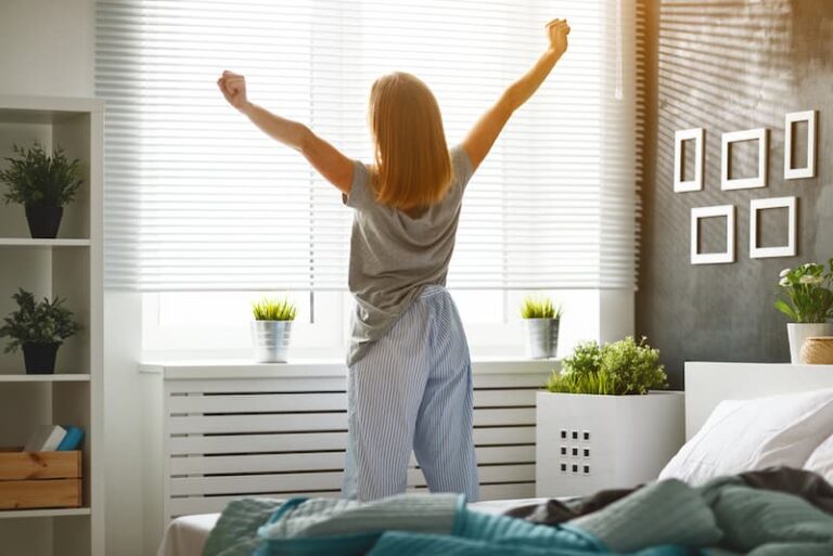 Eine Frau streckt sich am Fenster, wie kann man früh Aufstehen lernen?
