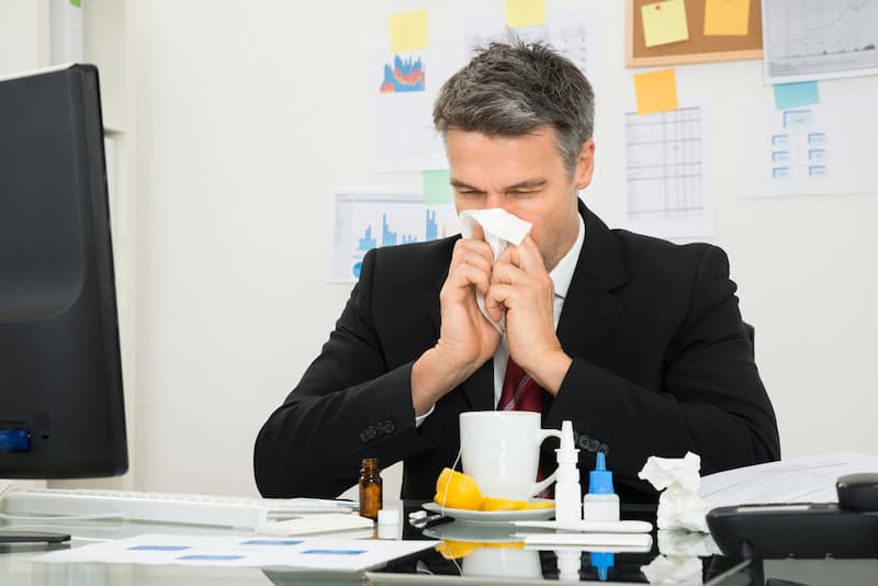 Ein Mann putzt sich die Nase am Arbeitsplatz, was ist Präsentismus?