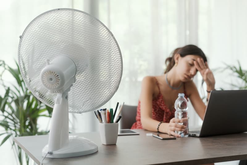 Eine Frau sitzt mit Ventilator im Büro, gibt es Hitzefrei im Job?