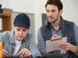 Ein angehender Elektroniker absolviert eine Externenprüfung. Sein Prüfer sitzt neben ihm und sieht dabei zu, wie er eine praktische Aufgabe bearbeitet.