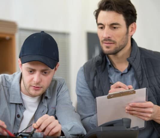Ein angehender Elektroniker absolviert eine Externenprüfung. Sein Prüfer sitzt neben ihm und sieht dabei zu, wie er eine praktische Aufgabe bearbeitet.