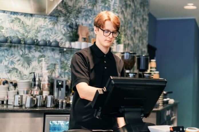 Ein Schüler steht im Bistro hinter der Kasse. Er übt einen Schülerjob aus.