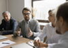 Ein Mitarbeiter sitzt am Tisch mit anderen Kollegen und Kolleginnen. Sie haben ein Meeting. Der Angestellte im Fokus möchte seine eigene Meinung vertreten und redet mit den anderen Meeting-Teilnehmenden, die ihm zuhören.