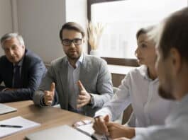 Ein Mitarbeiter sitzt am Tisch mit anderen Kollegen und Kolleginnen. Sie haben ein Meeting. Der Angestellte im Fokus möchte seine eigene Meinung vertreten und redet mit den anderen Meeting-Teilnehmenden, die ihm zuhören.