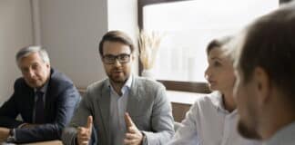 Ein Mitarbeiter sitzt am Tisch mit anderen Kollegen und Kolleginnen. Sie haben ein Meeting. Der Angestellte im Fokus möchte seine eigene Meinung vertreten und redet mit den anderen Meeting-Teilnehmenden, die ihm zuhören.