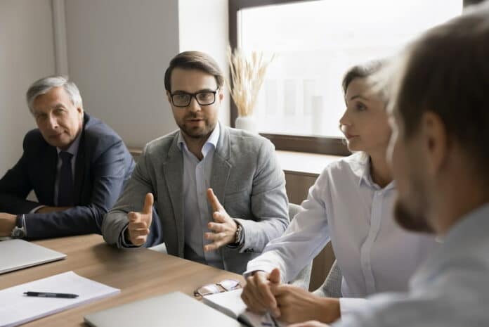 Ein Mitarbeiter sitzt am Tisch mit anderen Kollegen und Kolleginnen. Sie haben ein Meeting. Der Angestellte im Fokus möchte seine eigene Meinung vertreten und redet mit den anderen Meeting-Teilnehmenden, die ihm zuhören.
