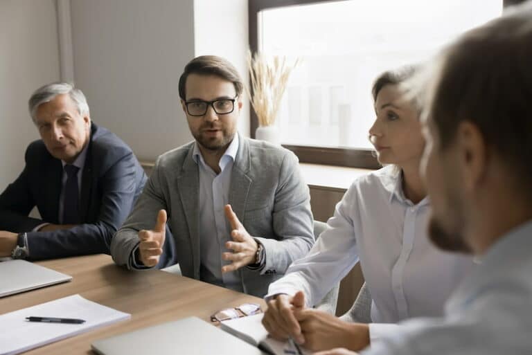 Ein Mitarbeiter sitzt am Tisch mit anderen Kollegen und Kolleginnen. Sie haben ein Meeting. Der Angestellte im Fokus möchte seine eigene Meinung vertreten und redet mit den anderen Meeting-Teilnehmenden, die ihm zuhören.
