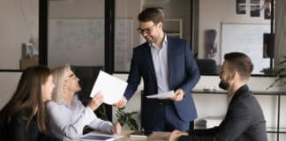 Arbeitnehmende sitzen in einem Büro an einem Tisch. Der Chef überreicht einer Arbeitnehmerin ein berufliches Zertifikat. Alle freuen sich über die Zertifizierung.