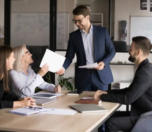 Arbeitnehmende sitzen in einem Büro an einem Tisch. Der Chef überreicht einer Arbeitnehmerin ein berufliches Zertifikat. Alle freuen sich über die Zertifizierung.