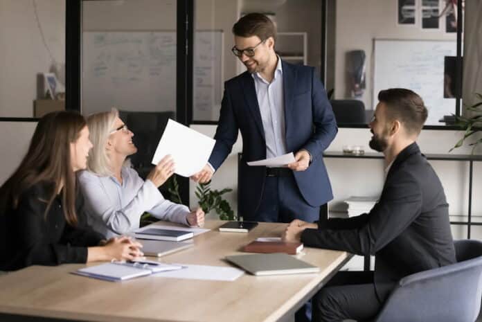Arbeitnehmende sitzen in einem Büro an einem Tisch. Der Chef überreicht einer Arbeitnehmerin ein berufliches Zertifikat. Alle freuen sich über die Zertifizierung.