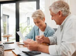 Ein Ehepaar gehobenen Alters sitzt an einem Tisch und berechnet mit dem Taschenrechner ihre private Altersvorsorge. Möglichkeiten, um vorzusorgen, lohnen sich. Mann und Frau lächeln.