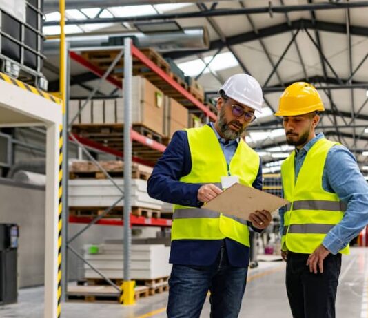 Ein Lagerarbeiter und ein Bewerber stehen in Warnweste in einem Lager. Der Bewerber erhält Erklärungen für sein Probearbeiten.