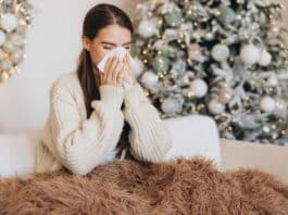 Eine Frau hat Schnupfen und sitzt mit einer Decke auf dem Sofa vor einem Weihnachtsbaum. Sie leidet in ihrem Weihnachtsurlaub unter Leisure Sickness.