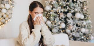 Eine Frau hat Schnupfen und sitzt mit einer Decke auf dem Sofa vor einem Weihnachtsbaum. Sie leidet in ihrem Weihnachtsurlaub unter Leisure Sickness.