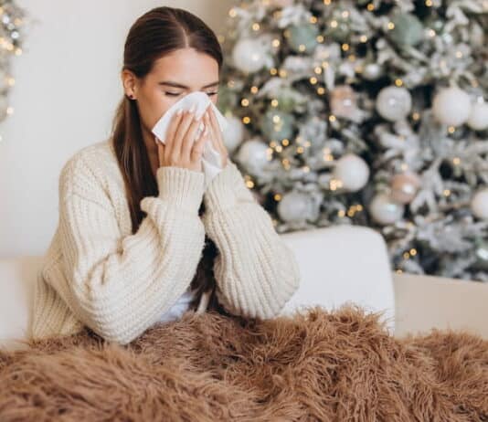 Eine Frau hat Schnupfen und sitzt mit einer Decke auf dem Sofa vor einem Weihnachtsbaum. Sie leidet in ihrem Weihnachtsurlaub unter Leisure Sickness.