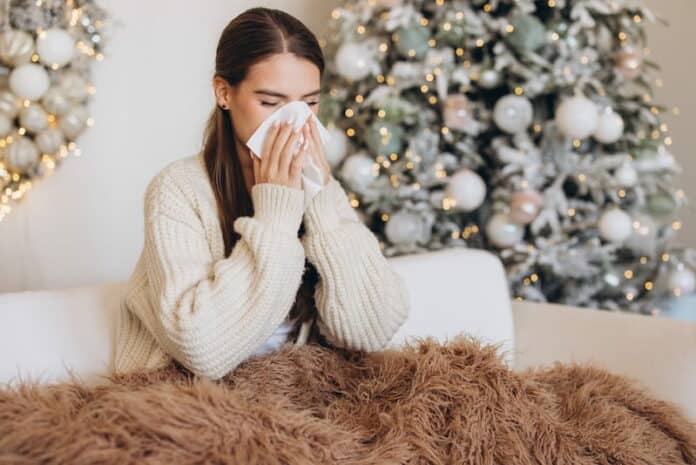 Eine Frau hat Schnupfen und sitzt mit einer Decke auf dem Sofa vor einem Weihnachtsbaum. Sie leidet in ihrem Weihnachtsurlaub unter Leisure Sickness.
