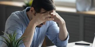 Mann sitzt mit den Händen im Gesicht am Schreibtisch. Er hat Angst vor der Arbeit.