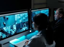 Zwei Sicherheitspersonen sitzen vor Monitoren, die Aufnahmen von Überwachungskameras zeigen, und sind in Arbeitsbereitschaft.