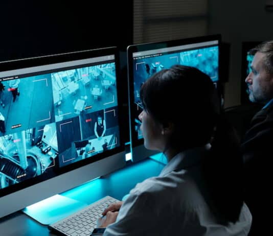 Zwei Sicherheitspersonen sitzen vor Monitoren, die Aufnahmen von Überwachungskameras zeigen, und sind in Arbeitsbereitschaft.
