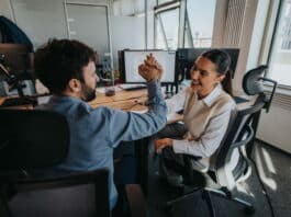 Zwei Beschäftigte klatschen sich am Arbeitsplatz in die Hände. Sie haben einen Erfolg erzielt und stärken ihre Selbstwirksamkeit im Beruf.