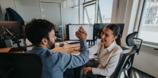 Zwei Beschäftigte klatschen sich am Arbeitsplatz in die Hände. Sie haben einen Erfolg erzielt und stärken ihre Selbstwirksamkeit im Beruf.