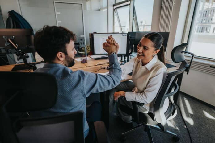 Zwei Beschäftigte klatschen sich am Arbeitsplatz in die Hände. Sie haben einen Erfolg erzielt und stärken ihre Selbstwirksamkeit im Beruf.